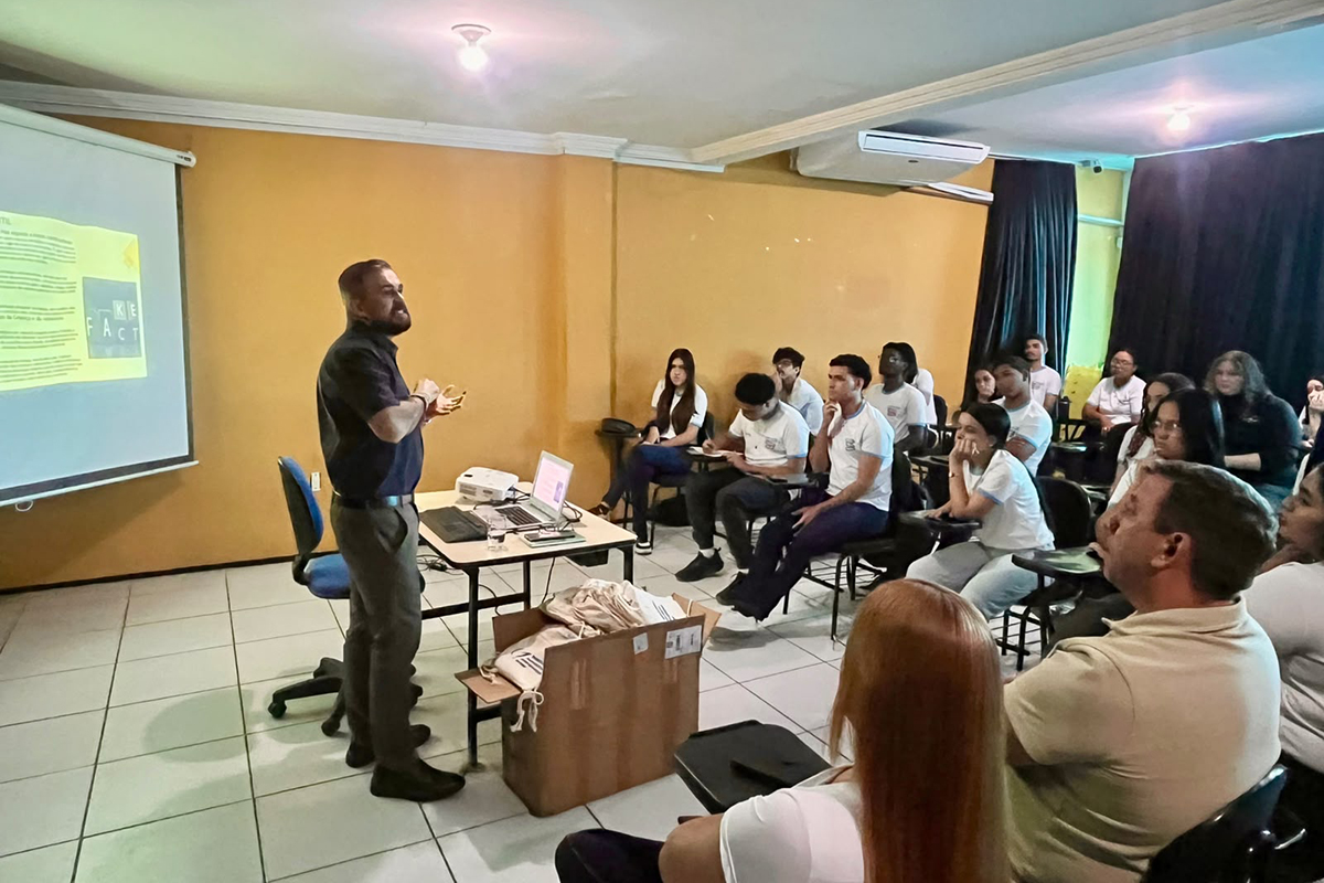 Em uma sala com paredes amarelas, um homem de barba e camisa escura gesticula de pé à esquerda enquanto ministra uma palestra para os jovens. A tela do projetor exibe um slide amarelo com o título "FAKE FACT", abordando o tema de notícias falsas. Vários jovens sentados em cadeiras escolares universitárias assistem à apresentação e fazem anotações em seus cadernos.