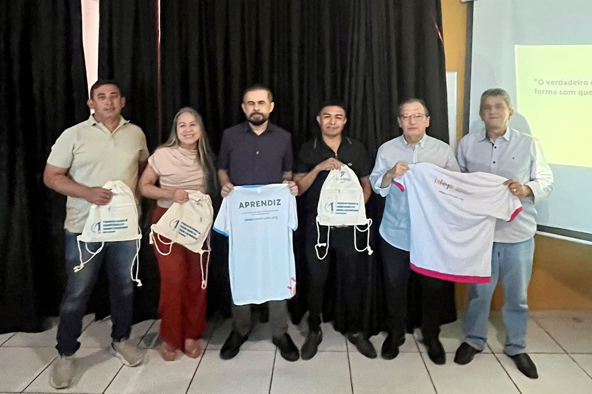 Seis adultos posam alinhados lado a lado e sorridentes à frente de uma cortina preta em um registro oficial de encerramento do evento. Eles seguram materiais institucionais do projeto, incluindo duas sacolas do tipo ecobag brancas com cordões e duas camisetas do programa. O homem de camisa escura no centro segura uma camiseta branca que traz a estampa "APRENDIZ".