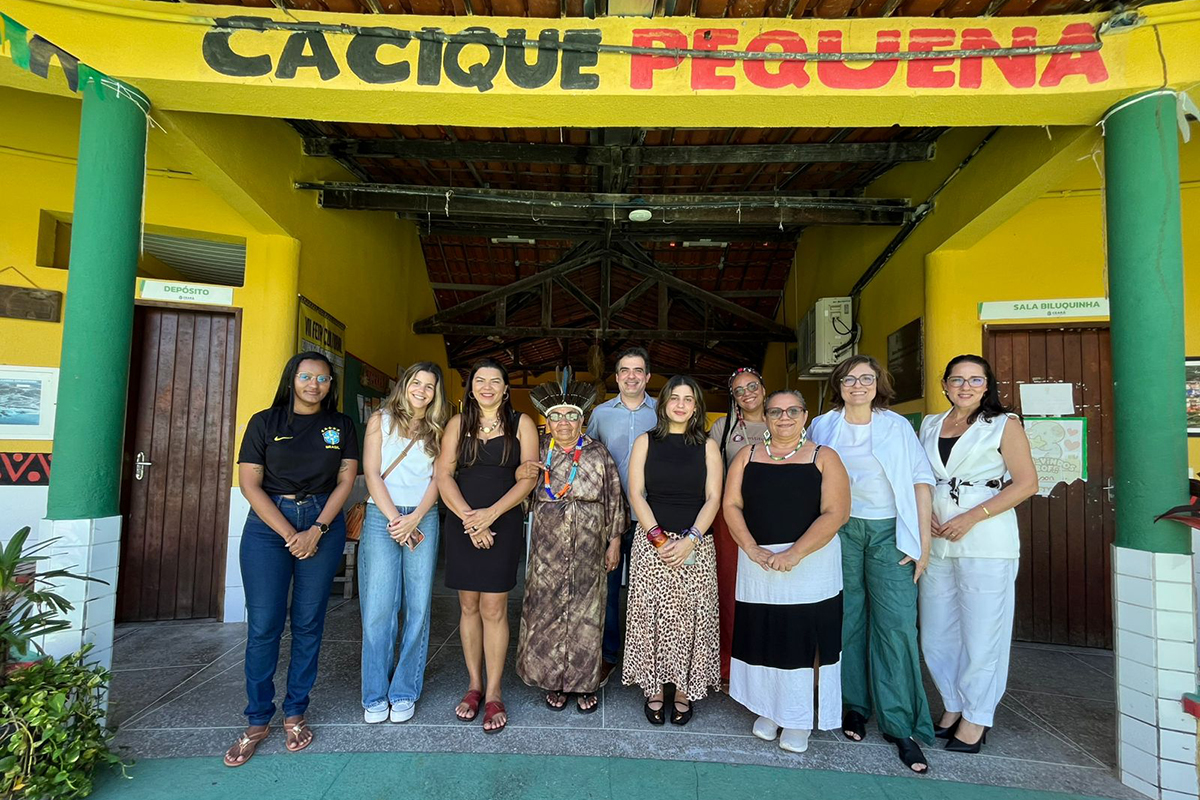Um grupo de dez pessoas está alinhado lado a lado na entrada de uma edificação amarela sob a inscrição "CACIQUE PEQUENA" pintada em letras pretas e vermelhas. No centro, destaca-se a mesma líder indígena idosa com seu cocar e colar colorido, ladeada por homens e mulheres em trajes casuais e formais. A estrutura possui colunas verdes e portas de madeira nas extremidades.