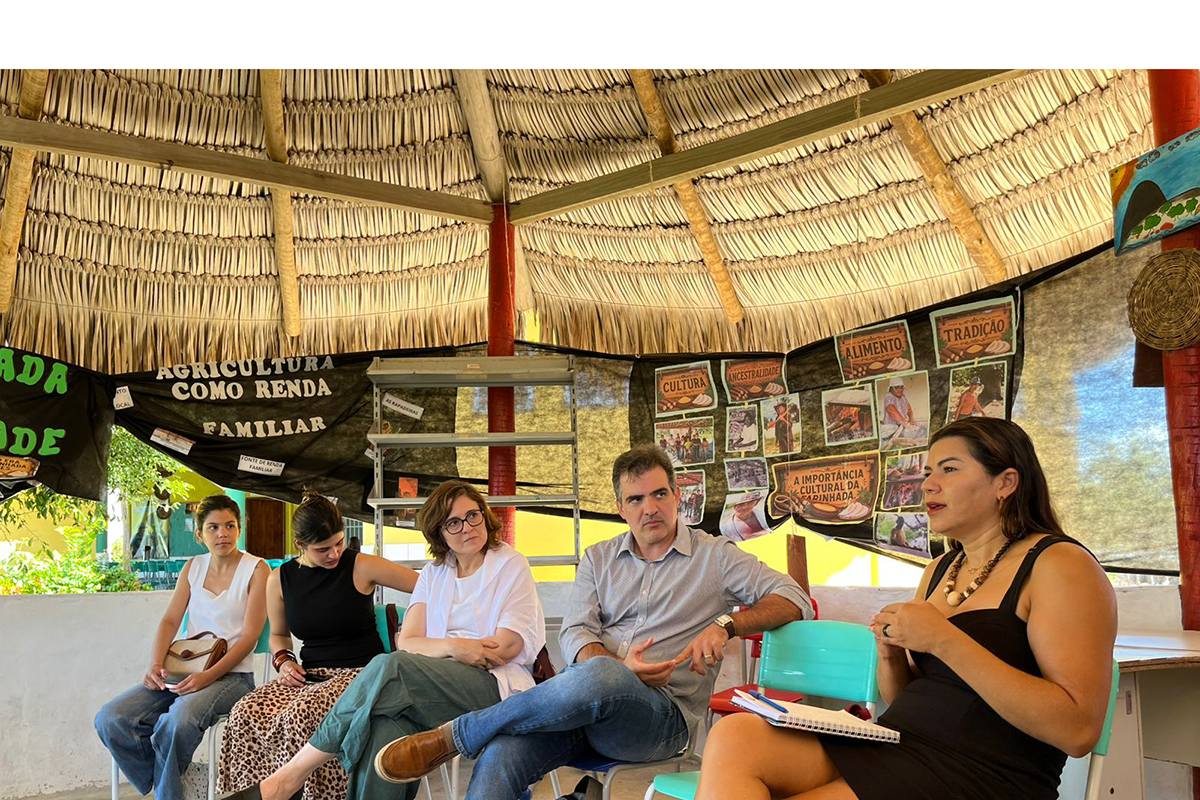 Cinco pessoas estão sentadas em semicírculo sob uma grande estrutura de teto de palha, prestando atenção a uma mulher em primeiro plano à direita que fala ativamente. Ao fundo, painéis pretos exibem fotos instrutivas e cartazes com títulos em letras garrafais como "AGRICULTURA COMO RENDA FAMILIAR", além de palavras como "CULTURA", "TRADIÇÃO" e "ALIMENTO".
