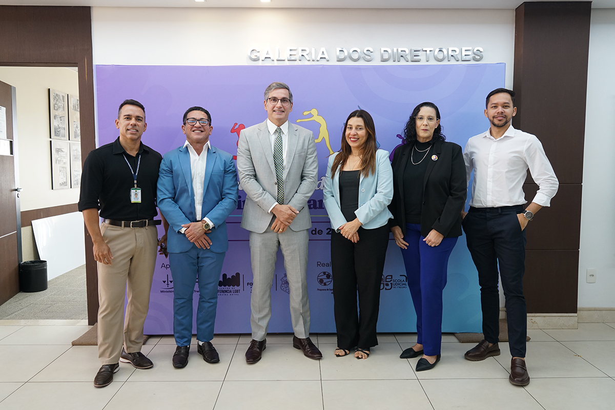 Seis pessoas posam alinhadas lado a lado em um ambiente interno, posicionadas à frente de uma estrutura com painel lilás e a inscrição "GALERIA DOS DIRETORES". Da esquerda para a direita, há três homens e duas mulheres vestindo trajes sociais e casuais finos, seguidos por mais um homem na ponta direita. Eles sorriem discretamente para a foto, mantendo as mãos unidas à frente do corpo ou posicionadas nos bolsos.