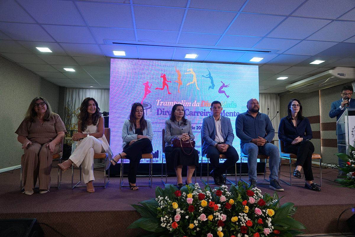Em um auditório, seis integrantes de uma mesa-redonda aparecem sentados em cadeiras alinhadas sobre um palco elevado, de frente para o público. Atrás deles, um grande painel de LED exibe o título "Trampolim da Diversidade - Direito, Carreira e Mente" com silhuetas coloridas em movimento. Na parte inferior, decorando a base do palco, destaca-se um grande e denso arranjo floral decorativo composto por rosas vermelhas, amarelas e brancas.