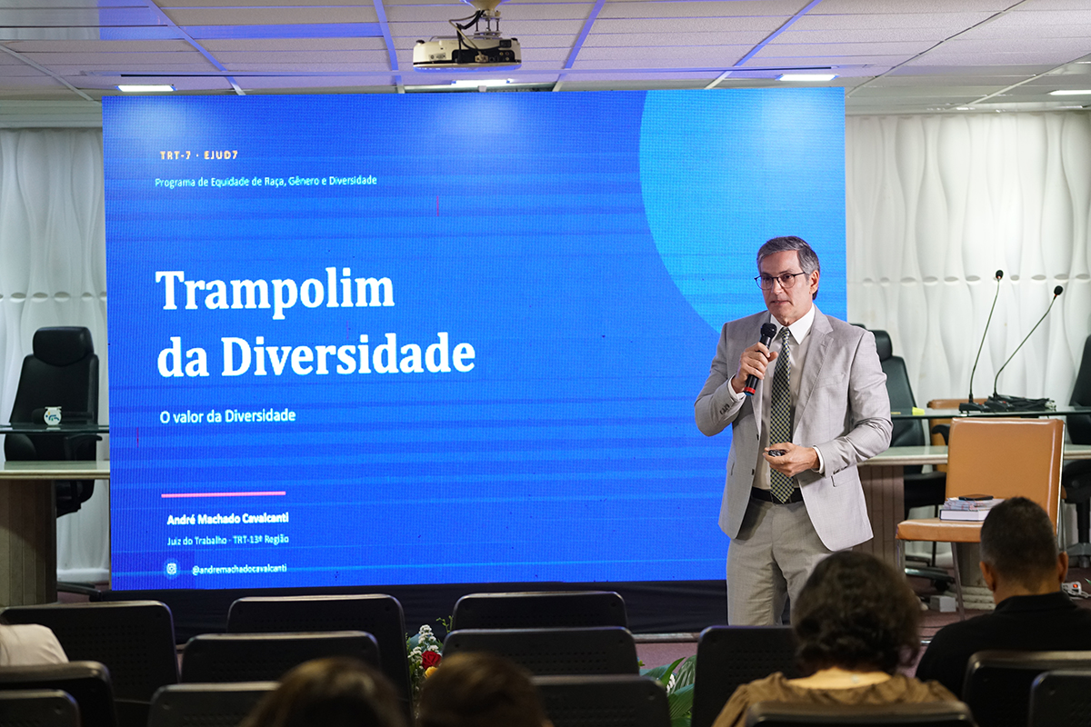 Um homem de óculos, vestindo terno cinza-claro, camisa branca e gravata, está em pé no lado direito do palco falando ao microfone. Ao fundo, um grande telão azul projeta em letras brancas o título da palestra: "Trampolim da Diversidade - O valor da Diversidade". Na parte inferior do enquadramento, em primeiro plano, aparecem as cabeças de alguns espectadores sentados na plateia de costas para a câmera.