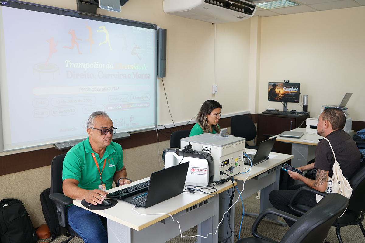 Em uma sala de apoio técnico ou escritório, um homem de camisa polo verde e óculos opera um notebook posicionado sobre uma mesa clara em primeiro plano. Ao fundo, uma mulher também de verde trabalha concentrada em outro computador portátil, enquanto um terceiro homem está sentado mais atrás olhando o celular. Na parede lateral esquerda, uma grande lousa digital exibe um slide promocional do evento "Trampolim da Diversidade".