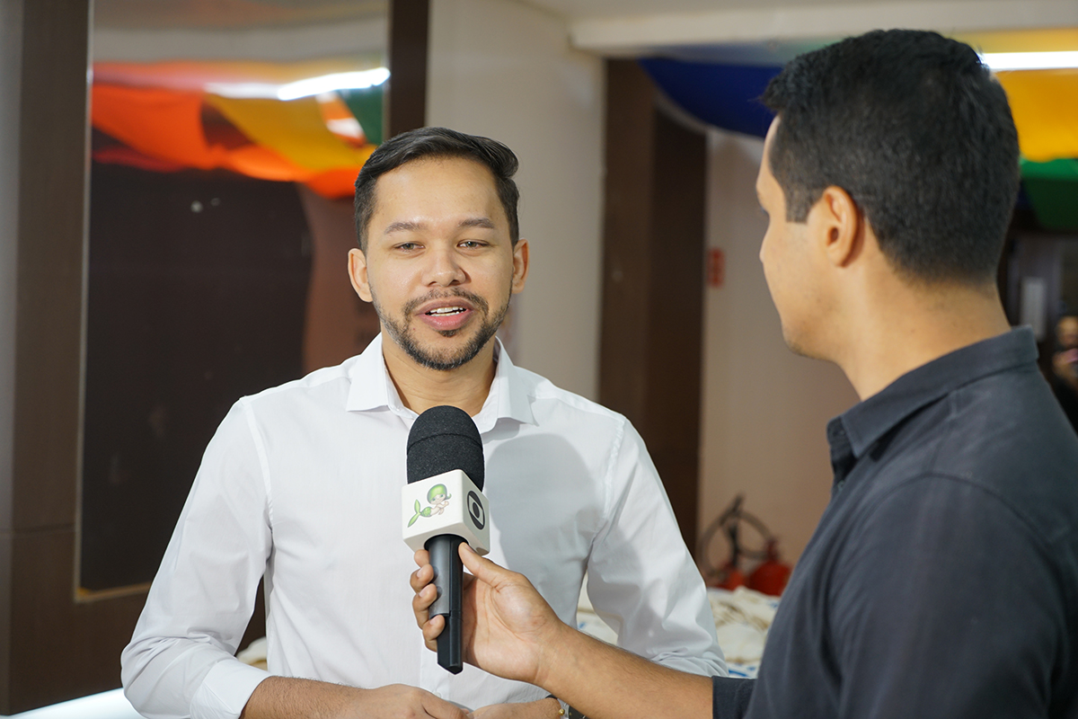 Um jovem de cabelos curtos, barba rala e camisa social branca aparece em plano médio focado enquanto concede uma entrevista. Uma mão em primeiro plano segura um microfone com canopla branca e logotipo verde em sua direção, captando a sua fala. O cenário de fundo é um ambiente interno de teto decorado com faixas coloridas nas cores do arco-íris, levemente desfocadas.