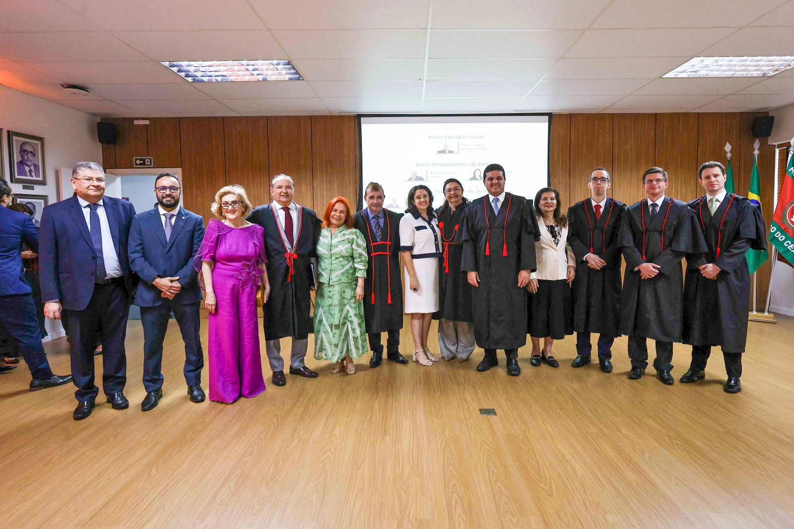 Fotografia em ambiente interno mostra treze pessoas alinhadas lado a lado em um auditório com piso de madeira e parede ao fundo em tom amadeirado. O grupo é composto por homens e mulheres vestindo trajes formais, sendo que vários deles usam togas pretas do Ministério Público com detalhes vermelhos.