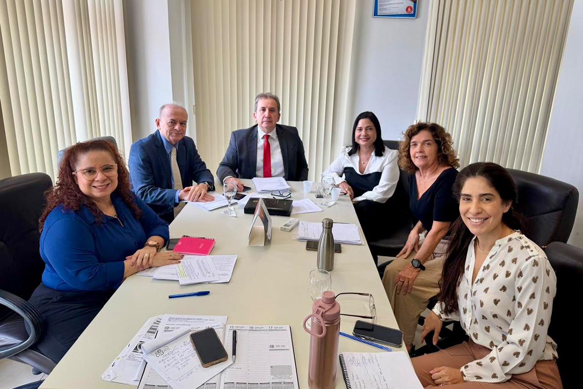 Fotografia em plano médio de uma reunião de trabalho em torno de uma mesa retangular branca. Seis pessoas estão dispostas ao redor da mesa: quatro mulheres e dois homens, todos sorrindo para a câmera em trajes esporte fino e formal. Sobre a mesa, há papéis organizados, cadernos, canetas, telefones celulares, copos e uma garrafa térmica rosa. O ambiente possui persianas verticais ao fundo e cadeiras de escritório giratórias na cor preta.