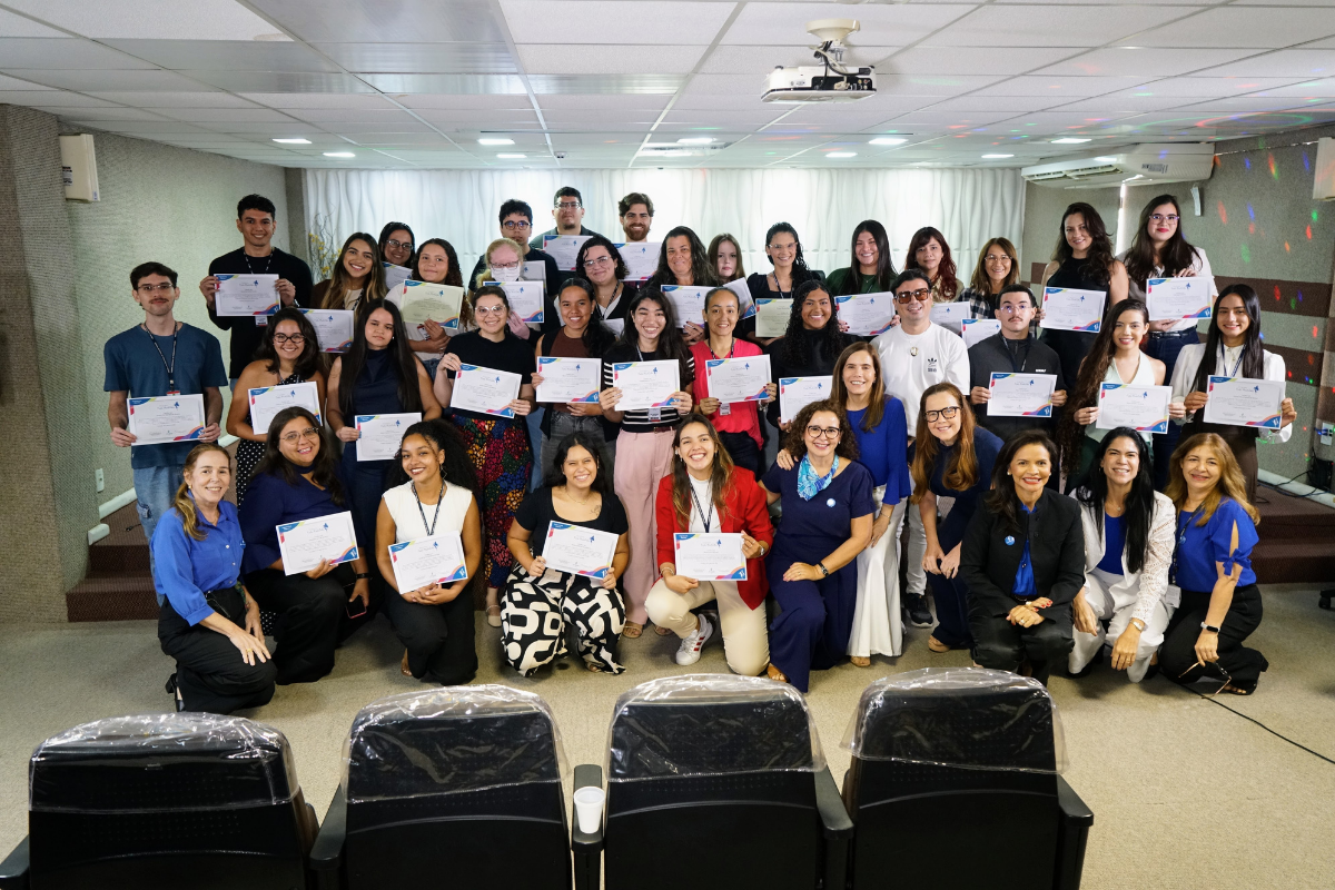 Fotografia mostra um grupo de aproximadamente 30 pessoas reunidas em uma sala, posando para a foto e segurando certificados de participação. Ao fundo, há uma parede com projeção de luzes coloridas, além de um projetor instalado no teto. Na parte frontal da imagem, aparecem algumas cadeiras de auditório vazias.