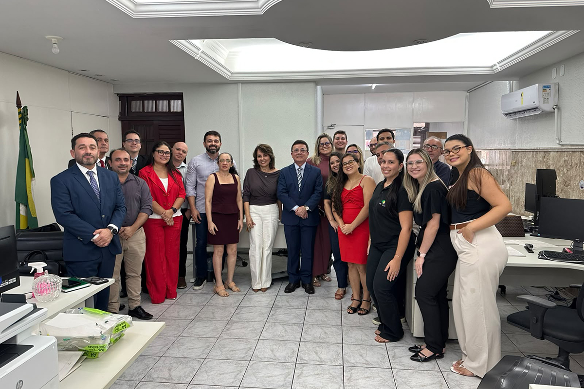 Fotografia em plano aberto e iluminação clara mostra um grupo de cerca de vinte pessoas reunidas e sorridentes no interior de um escritório bem iluminado, dispostas lado a lado e vestidas em trajes esporte fino e formais. No centro, destacam-se homens de terno e gravata e mulheres vestindo roupas em tons de vermelho, vinho e branco, enquanto ao fundo e nas laterais veem-se mesas de trabalho equipadas com computadores e uma bandeira do Brasil à esquerda.