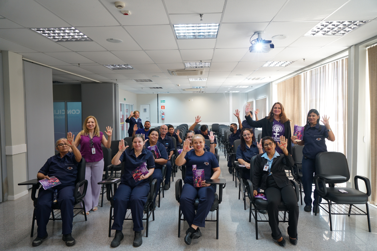 Cerca de 15 pessoas estão sentadas em cadeiras de estofado preto e base metálica preta, de frente para a câmera. Muitas pessoas vestem uniformes de cor azul marinho e quase todas as mulheres na foto mostram a palma da mão direita, sinalizando "pare". Outras pessoas seguram livros coloridos. A sala é ampla, com janelas de persianas no lado direito e um sistema de teto com luminárias e ar-condicionado.