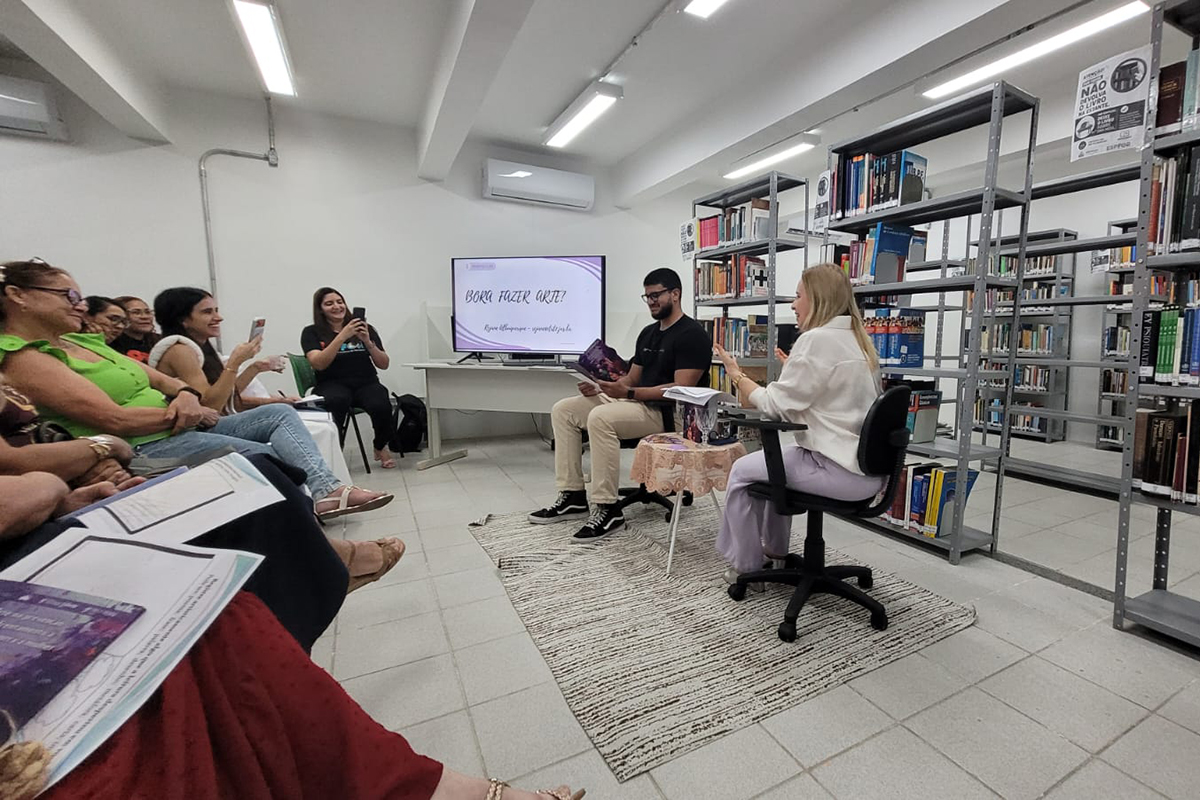 Em um ambiente de biblioteca com estantes metálicas cheias de livros, duas pessoas estão dispostas no centro sobre um tapete claro: um homem sentado em uma cadeira de escritório segurando um livro e uma mulher loira, de blusa branca e calça lilás, sentada em frente a ele falando. Ao lado esquerdo, parte da plateia assiste à apresentação sentada em fila, algumas pessoas registrando o momento com o celular, enquanto ao fundo há uma tela com o título "BORA FAZER ARTE?".