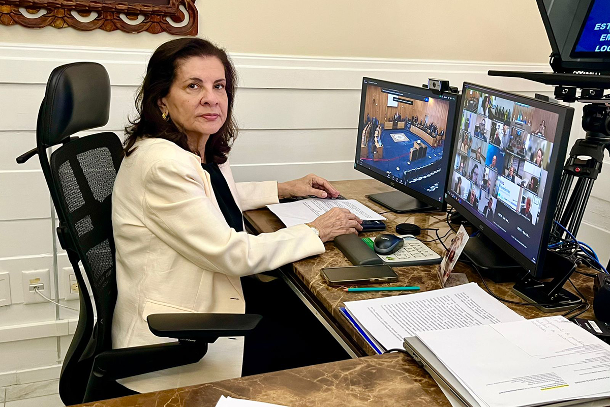 Mulher de pele clara e cabelos castanhos sentada em mesa de escritório, veste blazer off-white. Em sua frente, dois monitores exibem videoconferência com diversos participantes. Ela tem as mãos sobre um teclado e um mouse, com papéis impressos ao lado e um smartphone.