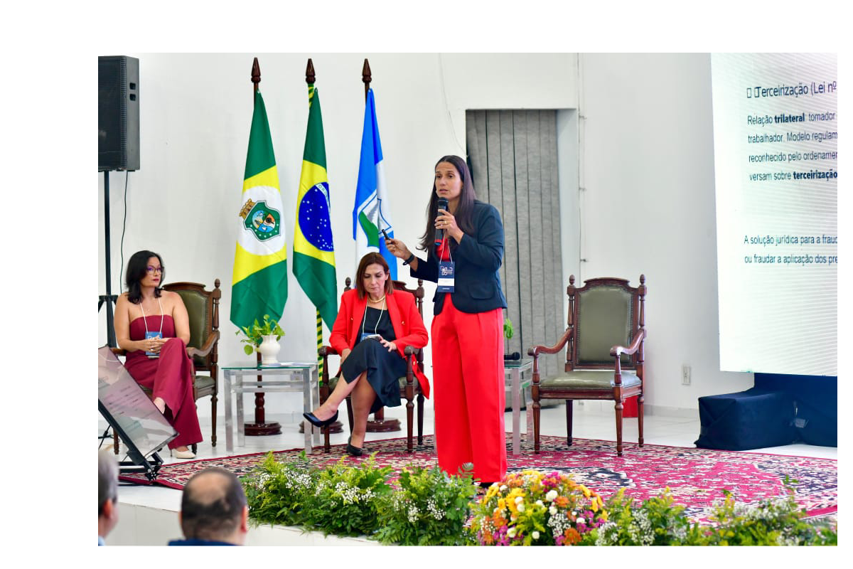 Fotografia em plano médio do palco do evento, onde uma mulher de cabelos longos, usando blazer escuro e calça vermelha, palestra em pé segurando um microfone e um passador de slides. À sua direita, projeta-se um telão com texto explicativo sobre terceirização, e ao fundo sobressaem-se três bandeiras oficiais dispostas entre duas mulheres sentadas em poltronas de madeira. A frente do palco é ornada com um grande tapete estampado e arranjos de plantas e flores coloridas.