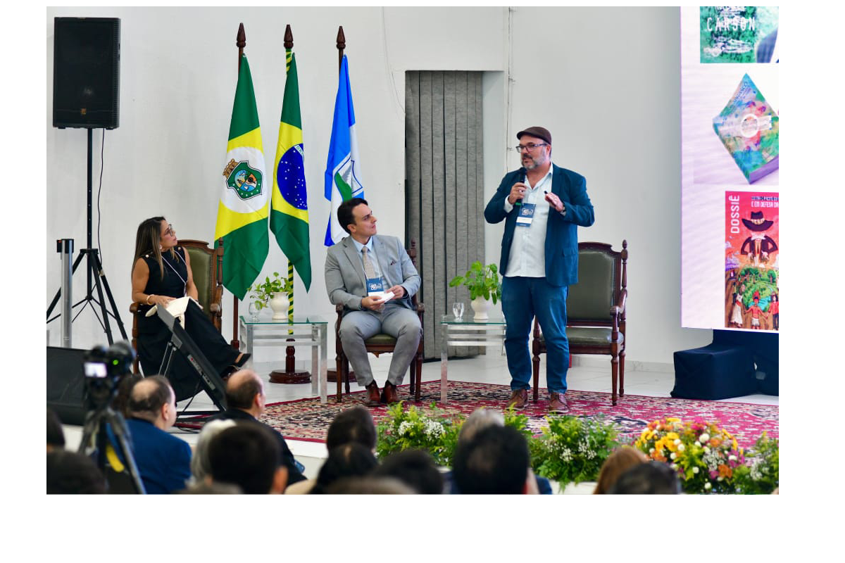 Fotografia em plano médio do palco do evento, com três bandeiras oficiais ao fundo (do município, do estado e do Brasil). À direita, um homem usando óculos, boina e blazer azul fala em pé ao microfone, enquanto ao seu lado, sentados em poltronas de madeira, uma mulher de vestido preto e um homem de terno cinza acompanham a exposição. Na borda direita, sobressai parte do telão com capas de publicações, e na parte inferior da imagem vê-se o público de costas.