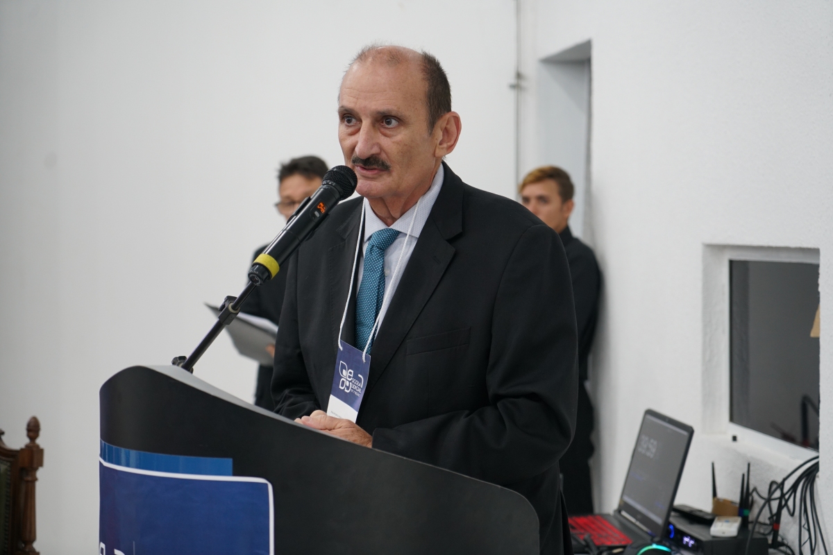 Fotografia em plano aproximado de um homem de meia-idade com bigode, vestindo terno preto, gravata azul-texturizada e crachá da Escola Judicial no pescoço. Ele está discursando de pé atrás de um púlpito escuro, falando diretamente ao microfone posicionado à sua frente. Ao fundo, contra paredes brancas, observam-se duas pessoas desfocadas e um notebook sobre uma bancada lateral.