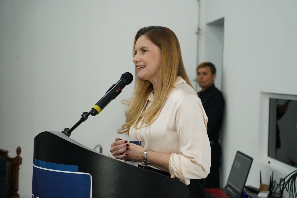Fotografia em plano médio aproximado de uma mulher loira de cabelos longos, vestindo uma camisa de cetim em tom claro e pulseira dourada no pulso. Ela está sorridente, discursando de pé atrás de um púlpito escuro com as mãos apoiadas à frente, falando em direção a um microfone fixado na haste. Ao fundo, sobressai a parede branca do ambiente com um homem vestindo camisa preta desfocado ao fundo.