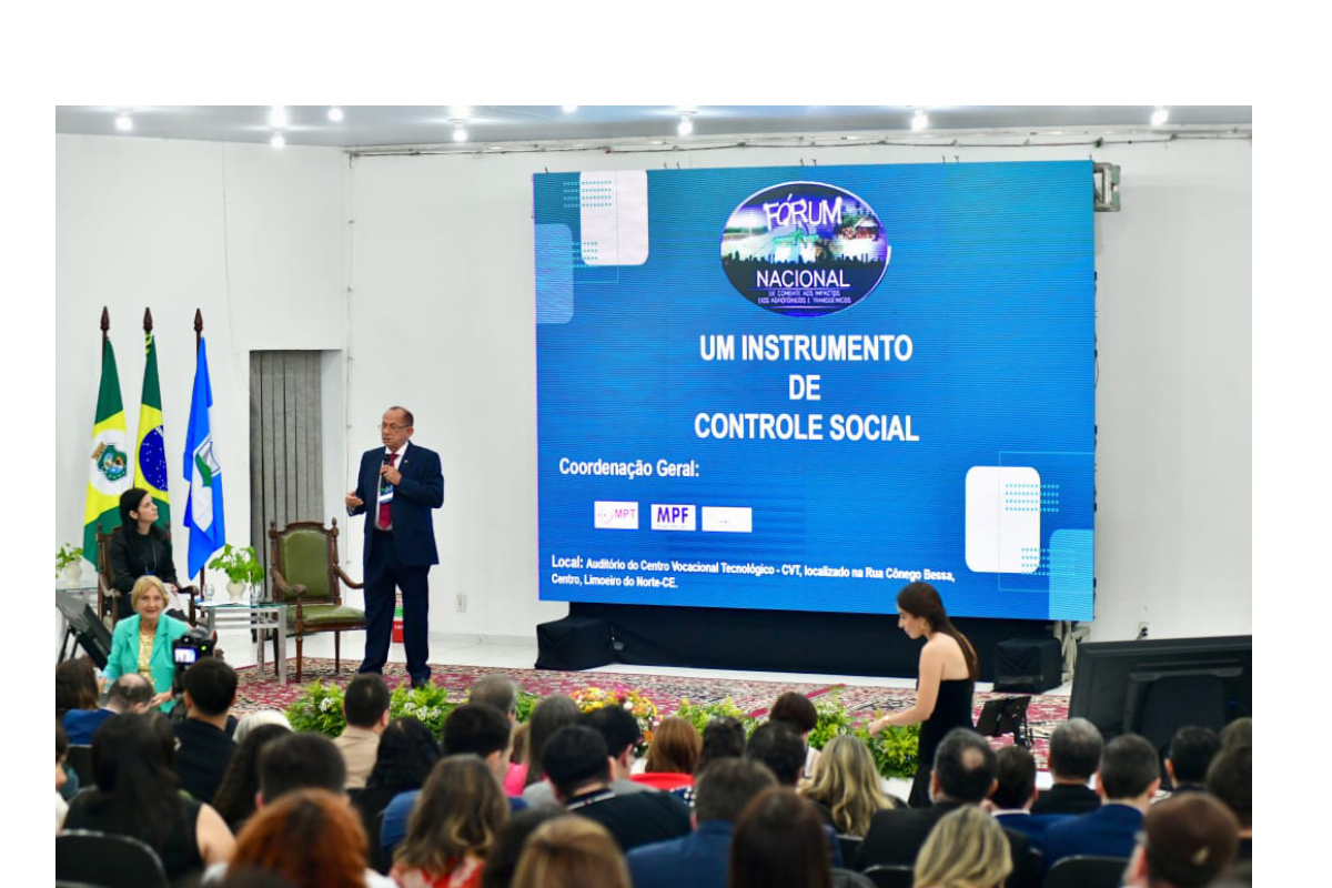 Fotografia em ângulo intermediário de um auditório lotado durante uma palestra. No palco, à esquerda, um palestrante homem de terno escuro e óculos fala em pé ao microfone, enquanto ao fundo um telão azul exibe a apresentação com o título "Um instrumento de controle social". À esquerda do palco, veem-se três bandeiras oficiais e outras integrantes da mesa sentadas, enquanto a plateia assiste atenta em primeiro plano.