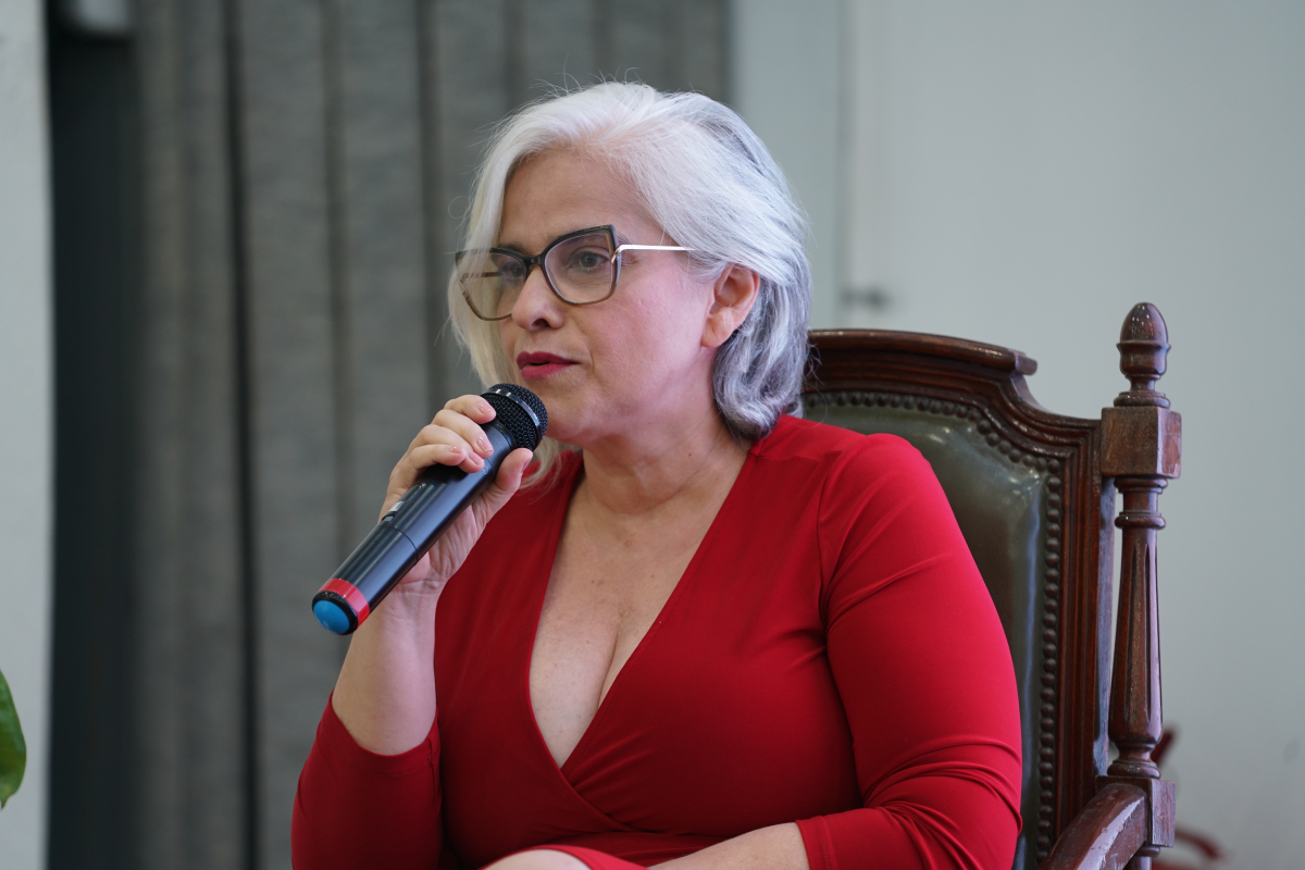 Fotografia em plano fechado de uma mulher de pele clara, cabelos grisalhos curtos e óculos de armação escura, vestindo um vestido vermelho de decote V. Ela está sentada em uma cadeira de madeira entalhada, segurando um microfone preto com a mão direita próximo ao rosto enquanto fala atentamente. Ao fundo, observa-se uma parede de tom neutro ao lado de uma cortina cinza em linhas verticais.