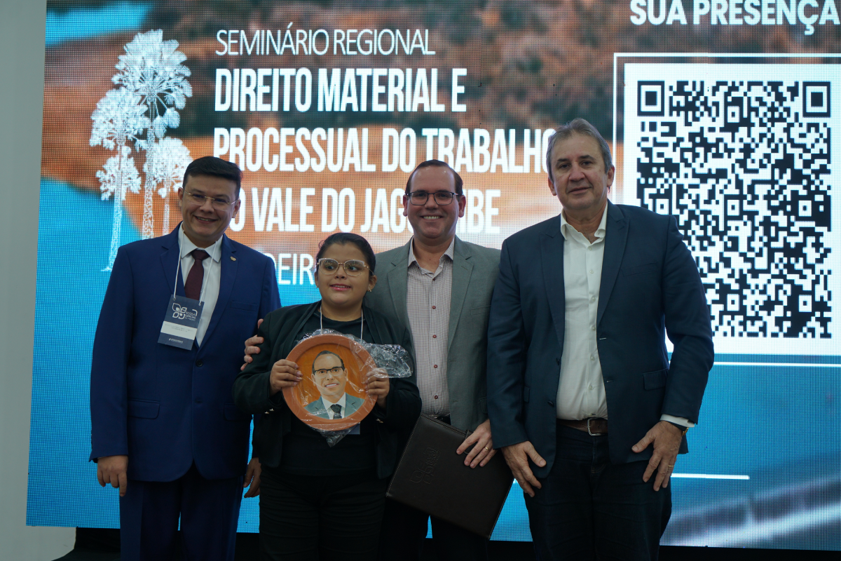Fotografia em plano médio de quatro pessoas lado a lado no palco, sorrindo em frente a um grande painel digital com QR code e a identificação do evento. No centro, uma jovem mulher segura um prato decorativo em cerâmica com o retrato pintado do palestrante posicionado ao seu lado. A composição é completada por dois homens de terno e gravata posicionados nas extremidades do grupo.