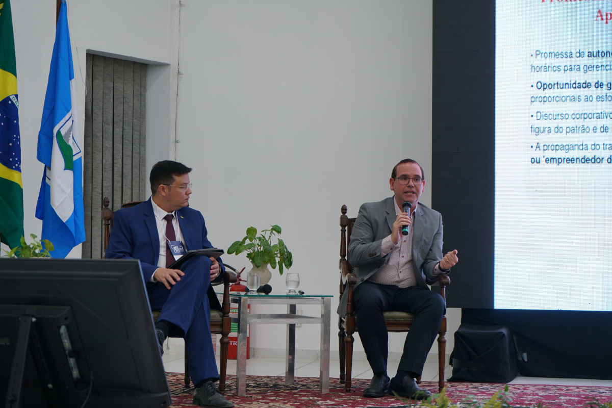 A imagem mostra um painel de discussão no palco, com dois homens brancos de meia-idade sentados em cadeiras de madeira e estofado marrom, em frente a um microfone e um monitor. À direita, um telão com o título "Promessa de autonomia", com quatro tópicos principais: "Promessa de autonomia e flexibilidade de horários para gerenciar os ganhos", "Oportunidade de ganho em taxas e bônus proporcionais ao esforço", "Discurso corporativo e colaboração, sem a figura de patrão e de chefe" e "A propaganda do trabalho autônomo ou empreendedorismo". À esquerda, três bandeiras: a bandeira do Brasil, a bandeira do Ceará e a bandeira do município. Há uma mesa de centro de vidro entre os palestrantes, com duas taças com água e um jarro com a planta jiboia. Há também um extintor de incêndio.