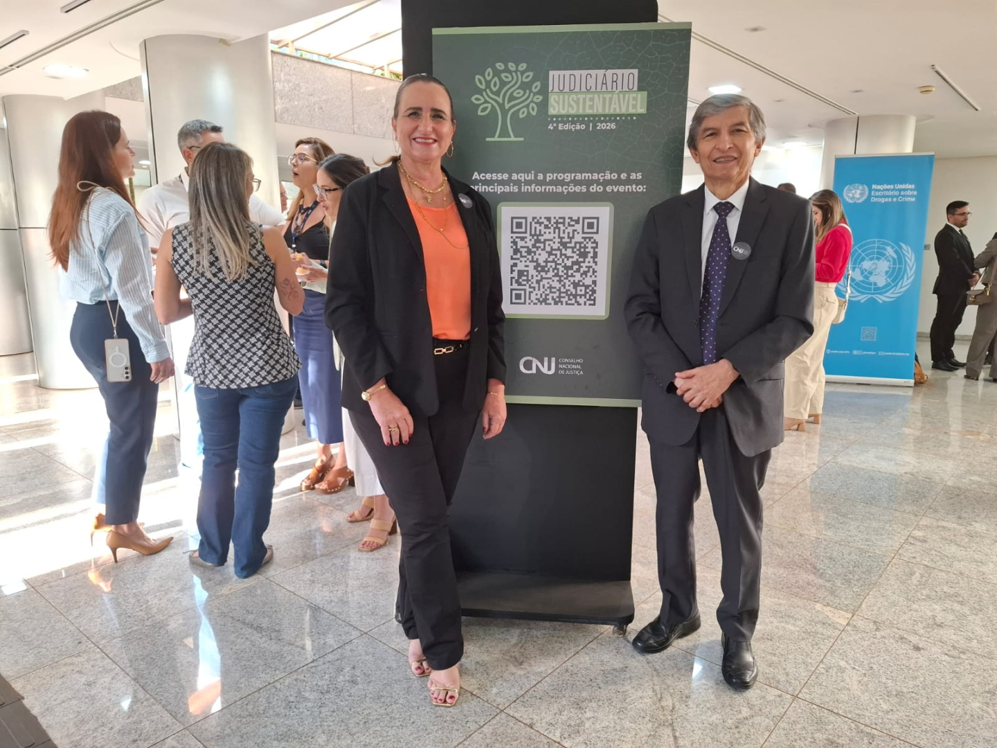 **Audiodescrição:** Fotografia colorida, em ambiente interno de um edifício institucional, com piso de granito e iluminação natural. Em primeiro plano, uma mulher e um homem estão lado a lado, em frente a um painel do evento “Judiciário Sustentável – 4ª Edição | 2026”, que apresenta um código QR e a marca do Conselho Nacional de Justiça (CNJ). A mulher veste blazer preto sobre blusa laranja, calça preta e sandálias, enquanto o homem usa terno cinza-escuro, camisa branca e gravata azul com estampas. Ao fundo, há outras pessoas conversando e um painel azul das Nações Unidas sobre Drogas e Crime.