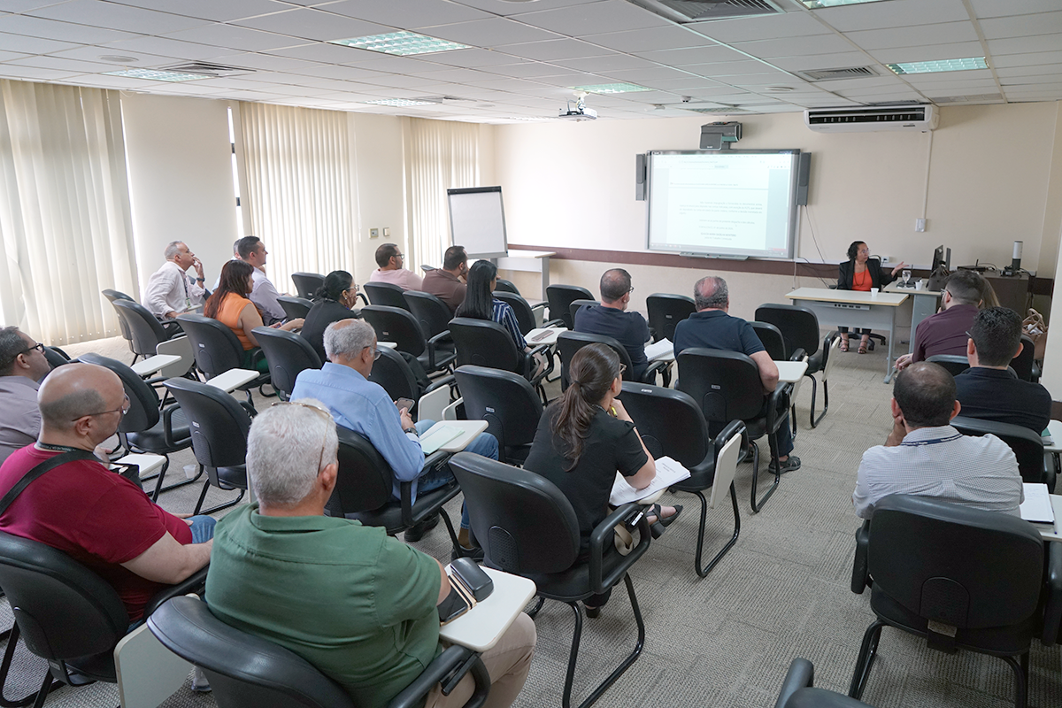 Fotografia em perspectiva de uma sala de aula de treinamento bem iluminada, vista por trás dos participantes sentados em carteiras estofadas escuras. Os alunos acompanham a apresentação voltados para a frente do ambiente, onde uma mulher de blazer preto e blusa laranja está sentada diante de uma mesa. À direita da instrutora, uma tela branca interativa exibe um texto projetado. Grandes janelas com persianas verticais claras cobrem a parede lateral esquerda.