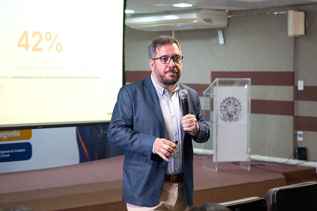 Foto em plano médio de um homem de cabelos e barba grisalhos, usando óculos de grau, camisa clara, blazer azul-marinho e calça bege. Ele está em pé, segurando um microfone na mão esquerda e um passador de slides na direita. Ao fundo, vê-se parte de uma tela de projeção com o número "42%" em destaque e um púlpito de acrílico transparente.