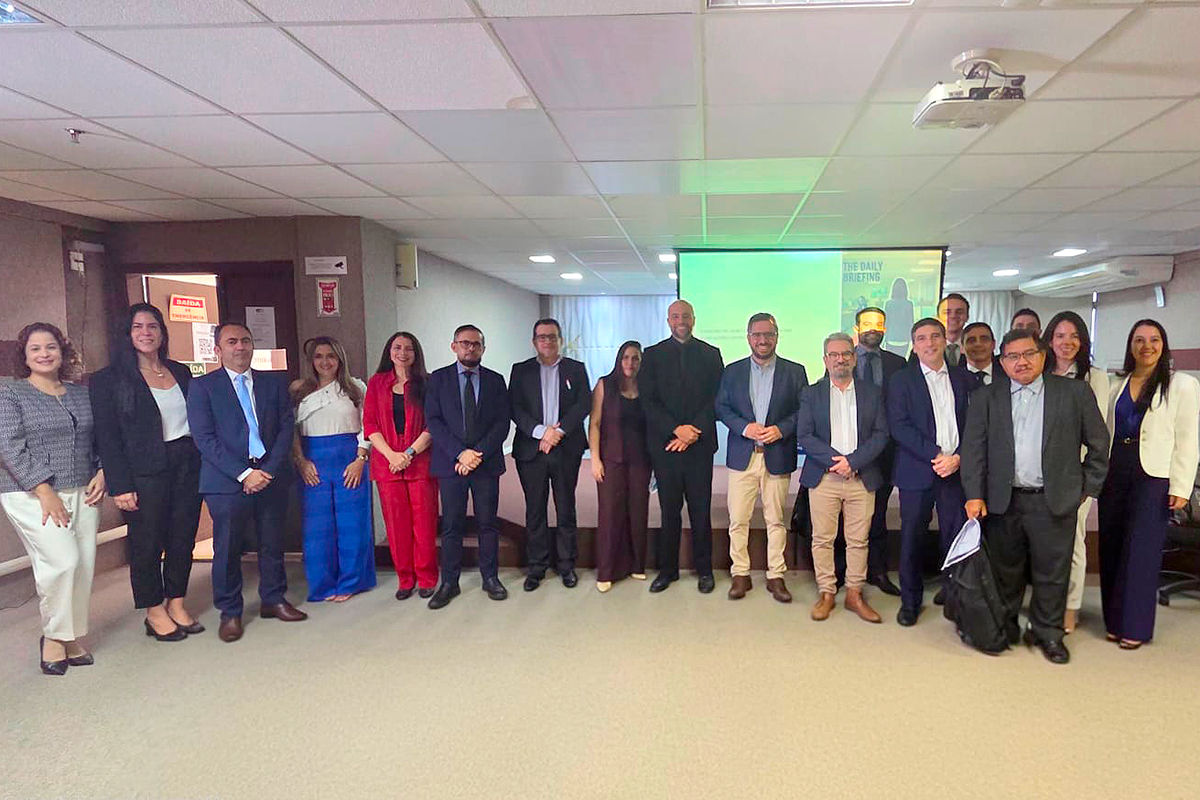 Foto em plano aberto mostra um grupo de cerca de vinte pessoas posadas lado a lado em um auditório. Elas usam trajes formais e esporte fino, como ternos, blazers e vestidos em tons escuros e coloridos. Ao fundo, no centro, há uma tela de projeção ligada, teto com luminárias embutidas e portas laterais de saída de emergência.