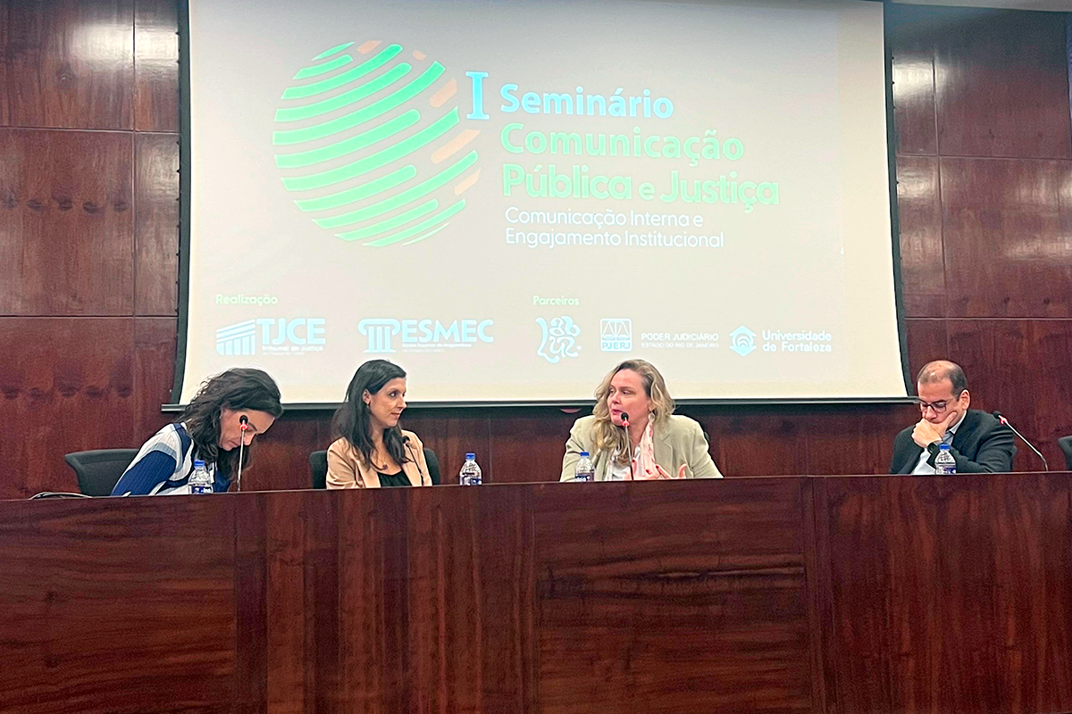 Quatro debatedores estão dispostos atrás de uma longa bancada de madeira em um auditório. No centro, uma mulher de blazer claro fala ao microfone, enquanto uma mulher à sua esquerda e um homem à sua direita escutam atentos; à ponta esquerda, outra palestrante consulta papéis. Acima deles, a projeção exibe a marca do evento com as logos dos organizadores e parceiros, como TJCE, ESMEC, PJERJ e Universidade de Fortaleza.