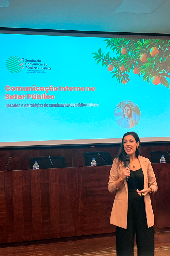 Uma mulher de cabelos escuros e blazer bege fala ao microfone em um auditório durante uma palestra. Ao fundo, uma grande tela exibe o título "I Seminário Comunicação Pública e Justiça: Comunicação Interna no Setor Público", decorada com folhagens e laranjas. A palestrante veste roupa preta por baixo do blazer e sorri enquanto se apresenta diante de uma bancada de madeira escura.