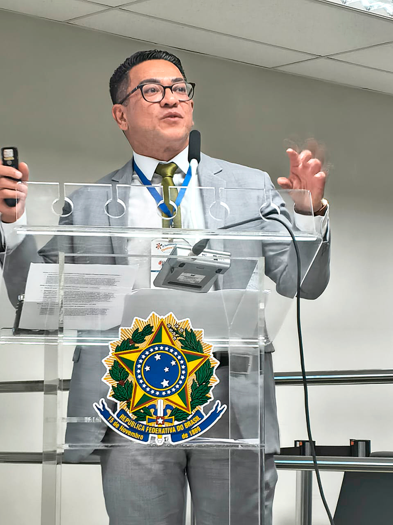Fotografia em plano médio fechado de homem de terno cinza e gravata verde discursando no púlpito de acrílico transparente. Ele usa óculos de armação preta, crachá no pescoço e segura um passador de slides na mão direita enquanto gesticula com a mão esquerda. O brasão das Armas do Brasil está destacado na parte frontal do púlpito.