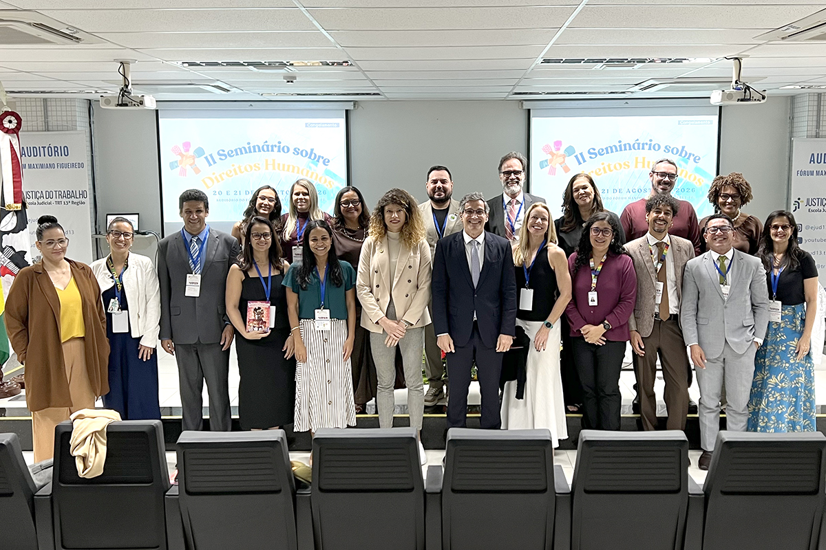 Fotografia em plano aberto de um grupo com cerca de dezenove pessoas alinhadas lado a lado em um auditório. Elas estão sorrindo para a câmera, vestindo trajes formais e casuais. Ao fundo, há duas telas de projeção com o texto "II Seminário sobre Direitos Humanos" e o brasão da Justiça do Trabalho. Na frente do grupo, veem-se as poltronas pretas do auditório.