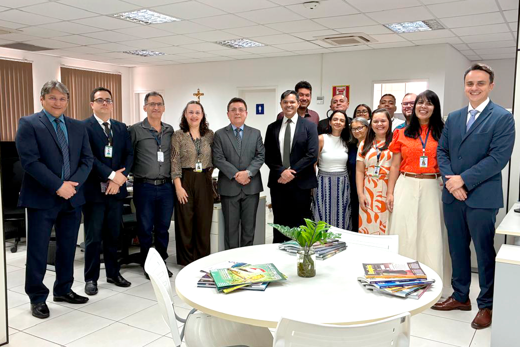 Fotografia em ambiente corporativo com cerca de quinze pessoas posadas lado a lado em semicírculo, sorrindo para a câmera. Homens e mulheres vestem trajes formais e esporte fino, como ternos, gravatas, camisas sociais e vestidos coloridos, alguns usando crachá. O local é um escritório amplo e iluminado, com paredes brancas, piso claro, janelas com persianas ao fundo e teto modular com luminárias fluorescentes. Em primeiro plano, destaca-se uma mesa redonda branca com revistas organizadas sobre o tampo e um pequeno vaso com folhagem no centro.