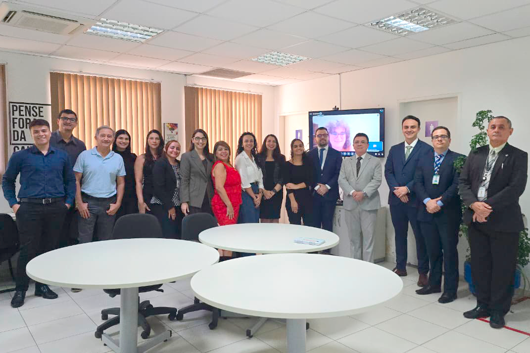Fotografia em ambiente interno e bem iluminado reúne cerca de 15 pessoas alinhadas lado a lado em pé, sorrindo para a câmera. O grupo veste trajes formais e esporte fino, com homens de terno ou camisa social e mulheres em terninhos, vestidos e blusas elegantes. Em primeiro plano, há três mesas redondas brancas em uma sala de escritório com paredes claras e persianas ao fundo. Na parede ao fundo, uma tela de televisão exibe a videochamada de uma mulher, e à direita há uma planta decorativa.