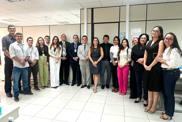 Fotografia colorida em plano aberto mostrando um grupo de dezessete pessoas alinhadas em um escritório bem iluminado. Homens e mulheres vestem trajes formais e esporte fino, posicionados lado a lado sorrindo para a câmera. Ao fundo, há divisórias de escritório beges, teto modular com luminárias embutidas e piso de cerâmica clara.