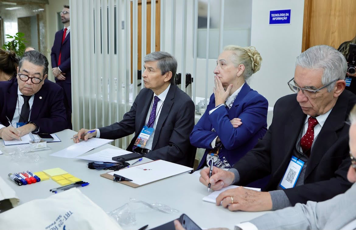 Quatro participantes de um evento institucional estão sentados em uma mesa de reunião, prestando atenção à apresentação. Eles vestem ternos e trajes formais, munidos de papéis, canetas e crachás de identificação do evento sobre a mesa. Ao fundo, observa-se a estrutura interna de um escritório com divisórias e sinalização de ambiente.