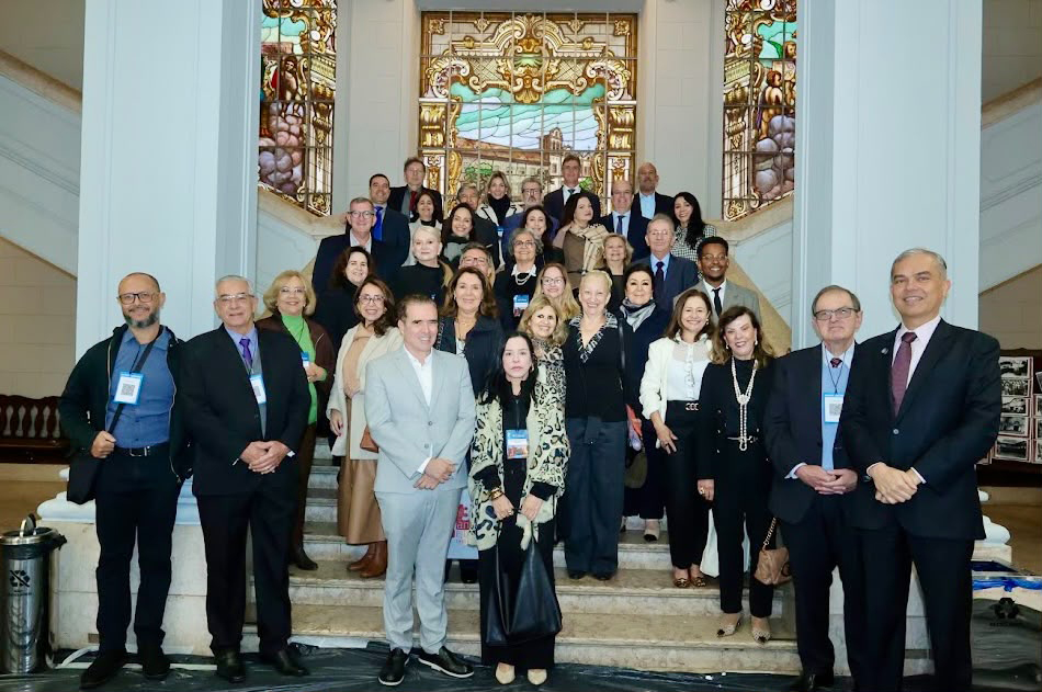Um grande grupo de pessoas posa para uma foto coletiva nos degraus de uma imponente escadaria de mármore. Ao fundo sobressai um detalhado painel de vitral colorido emoldurado por colunas e elementos arquitetônicos clássicos. Todos vestem roupas sociais e elegantes, dispostos em vários níveis ao longo dos degraus da escada.
