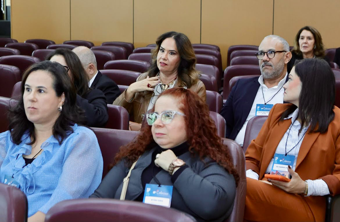 Visão em plano médio de auditório mostrando participantes sentados em poltronas estofadas durante uma palestra ou reunião. Em destaque no primeiro plano, uma mulher com óculos de armação clara e cabelos cacheados olha atentamente para a frente. Os demais presentes, vestidos com trajes formais e portando crachás, acompanham a sessão com expressões concentradas.