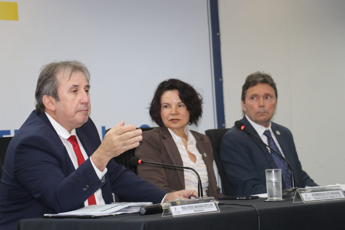 Três autoridades do Judiciário estão sentadas à mesa de evento formal vestindo trajes sociais. Em primeiro plano, o conselheiro Paulo Régis gesticula com a mão esquerda enquanto fala. Ao fundo, um painel institucional exibe parte da identidade visual da Justiça do Trabalho.