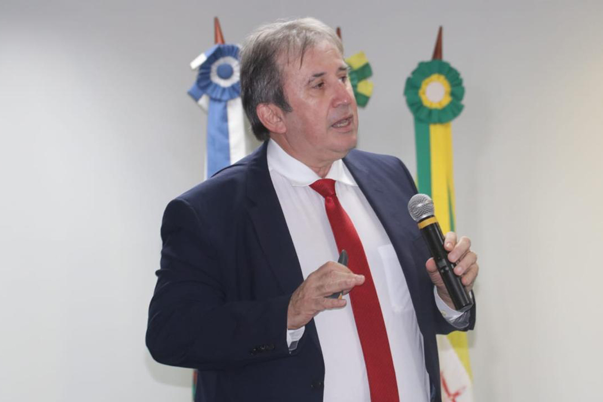 O conselheiro Paulo Régis é fotografado em plano médio vestindo terno azul e gravata vermelha. Ele segura um microfone com a mão esquerda enquanto fala e gesticula levemente com a direita. Atrás dele, ao fundo, sobressaem-se três bandeiras oficiais dispostas em mastros.