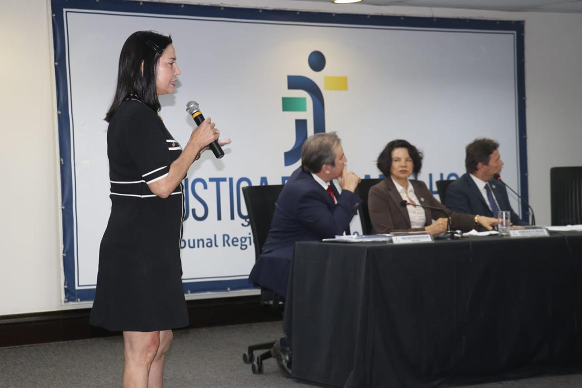 Uma mulher de vestido preto de mangas curtas fala em pé segurando um microfone à esquerda. Três integrantes da mesa diretora escutam atentamente a apresentação voltados para ela. Ao fundo, destaca-se um grande painel com a marca e a inscrição da Justiça do Trabalho.