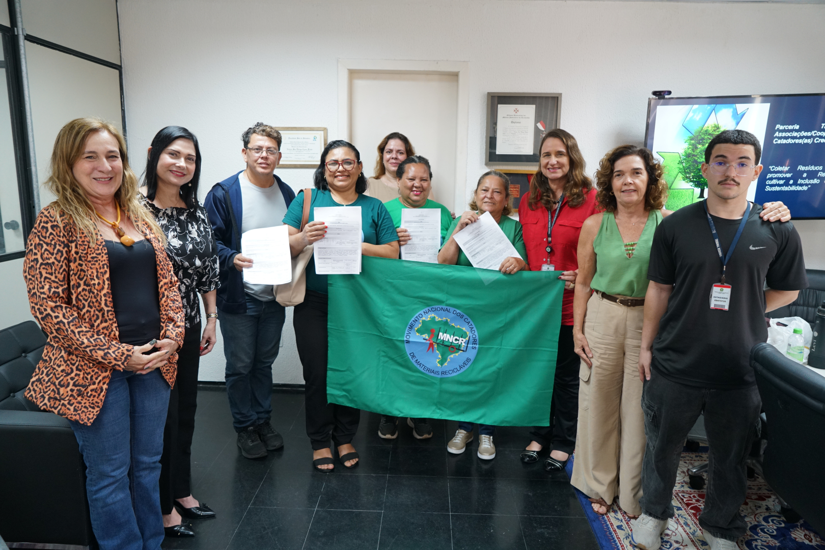A fotografia mostra um grupo de nove pessoas reunidas em uma sala, posando para a câmera. Ao centro, algumas pessoas seguram documentos e uma grande bandeira verde do Movimento Nacional dos Catadores de Materiais Recicláveis (MNCR). Ao fundo, há uma televisão exibindo uma apresentação e, nas paredes, quadros e certificados.