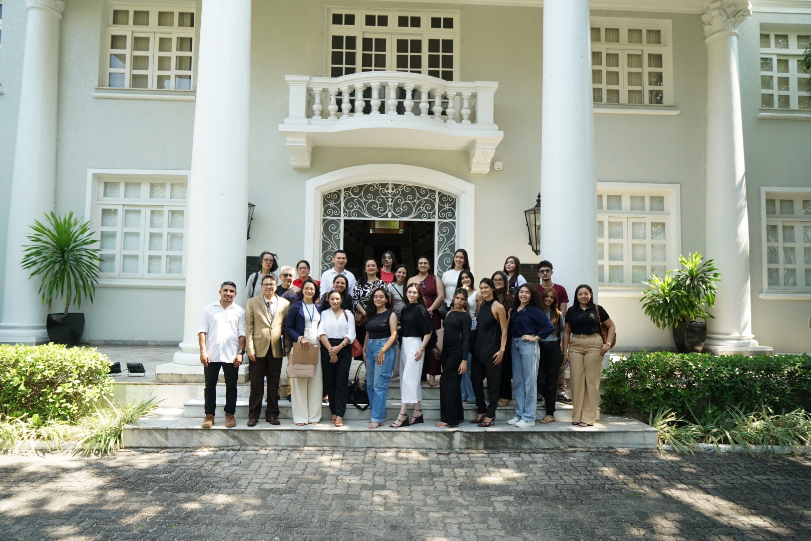 Fotografia em plano aberto mostra um grupo de aproximadamente 30 pessoas reunidas em frente à entrada de um prédio institucional. Os participantes estão posicionados sobre uma escadaria, em diferentes poses, olhando para a câmera. Ao fundo, destaca-se a fachada clara do edifício, com grandes colunas brancas, janelas e uma porta central ornamentada com detalhes em ferro. Há jardins com plantas verdes em ambos os lados da escadaria. A imagem registra um momento coletivo, provavelmente relacionado a uma visita ou atividade institucional.