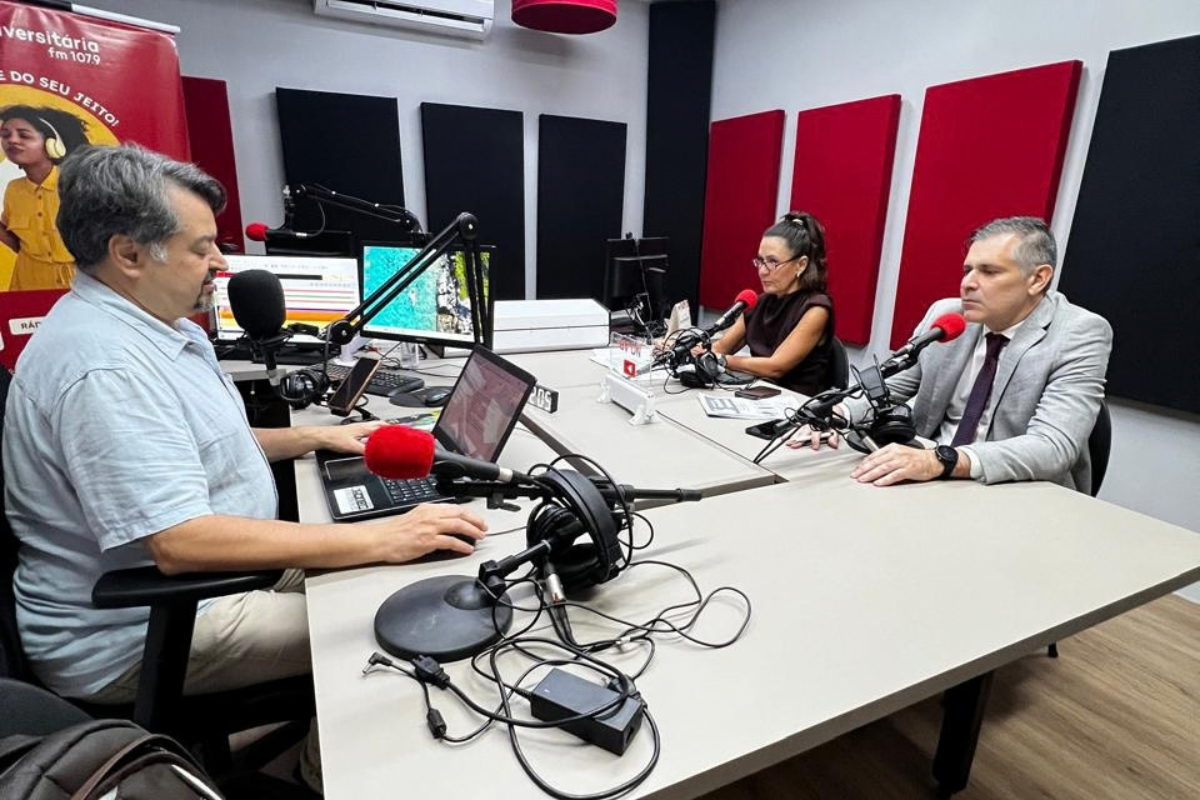 Em um estúdio de rádio, três pessoas estão sentadas ao redor de uma mesa equipada com microfones, fones de ouvido e computadores. À esquerda, um homem opera os equipamentos enquanto conversa com os outros dois participantes. Ao centro, uma mulher usa fones de ouvido diante do microfone. À direita, um homem de terno e gravata participa da conversa. Ao fundo, há painéis acústicos nas cores preta e vermelha.