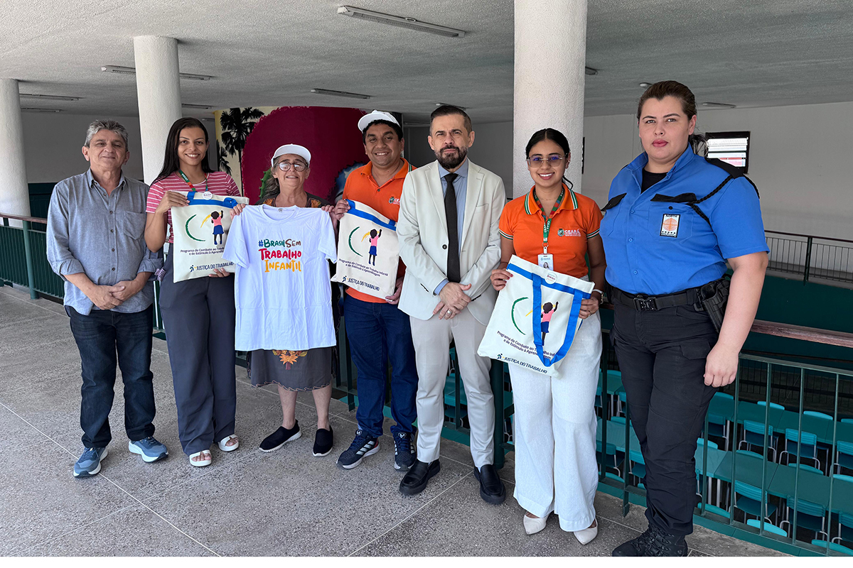 Sete pessoas posam lado a lado em um corredor coberto com pilares brancos e parapeito verde. No centro, o magistrado veste terno cinza claro ao lado de educadores que seguram sacolas de pano e uma camisa branca com a frase "#BrasilSemTrabalhoInfantil". À direita, uma profissional de farda azul-marinho completa o grupo.