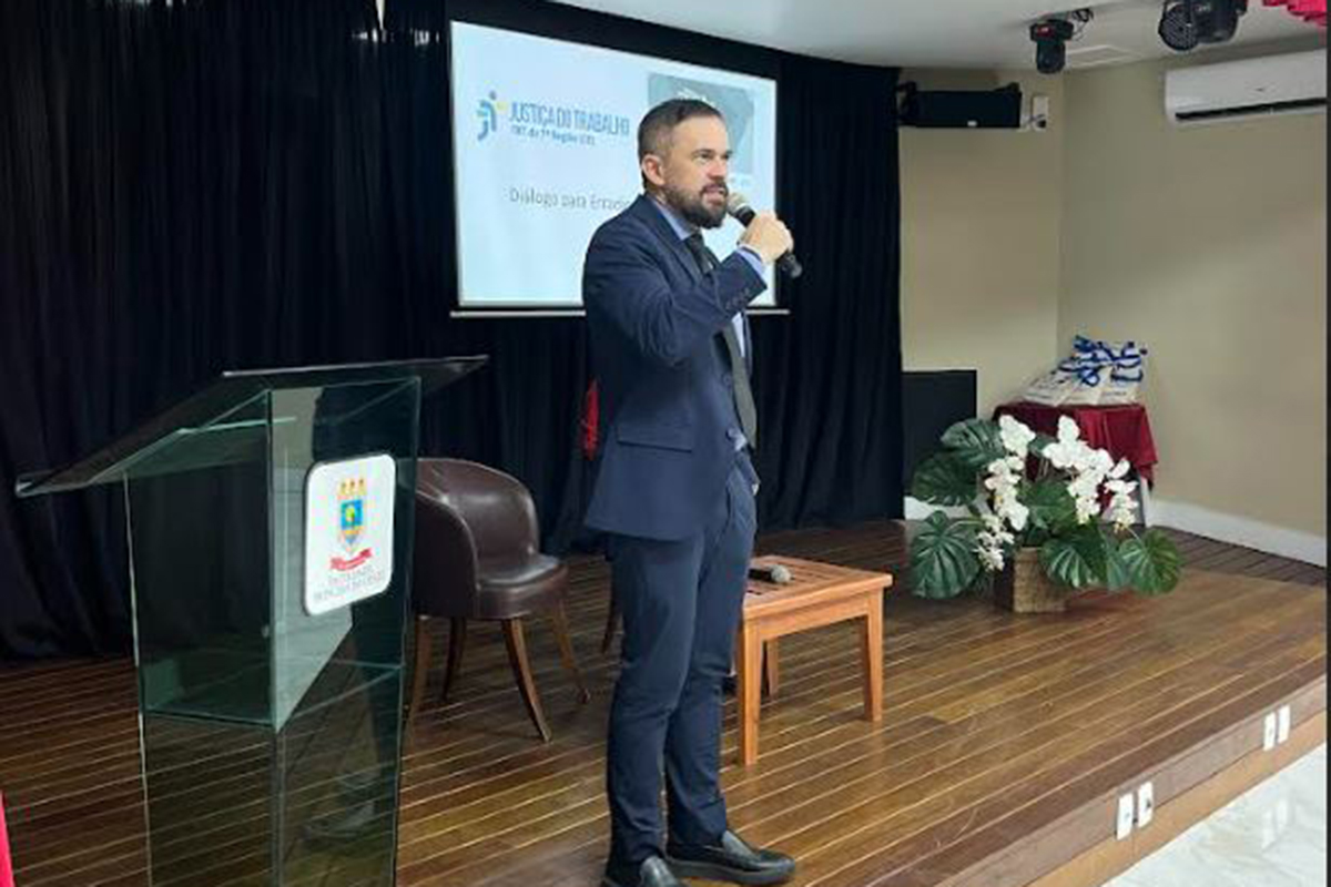 Em palco com piso de madeira, o juiz Célio Timbó fala em pé segurando um microfone na mão direita. Ele veste terno azul-marinho sobre camisa social e gravata escura. Ao fundo, uma tela de projeção exibe a apresentação do evento ao lado de um púlpito de acrílico e vasos com plantas decorativas.
