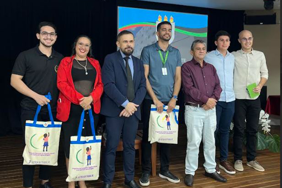 Sete pessoas posam alinhadas no palco da faculdade em frente ao painel do evento. O juiz de terno escuro ocupa a posição central acompanhado por professores e gestores da instituição. Três dos participantes seguram sacolas institucionais brancas e azuis com a ilustração do programa estampada.