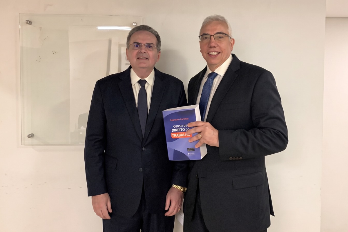 Na imagem, dois homens aparecem lado a lado, ambos usando terno, camisa social e gravata. O homem à direita segura um livro de Direito do Trabalho e sorri para a câmera. O homem à esquerda também olha para a câmera, com expressão séria. Eles estão em um ambiente interno, com uma parede clara ao fundo.