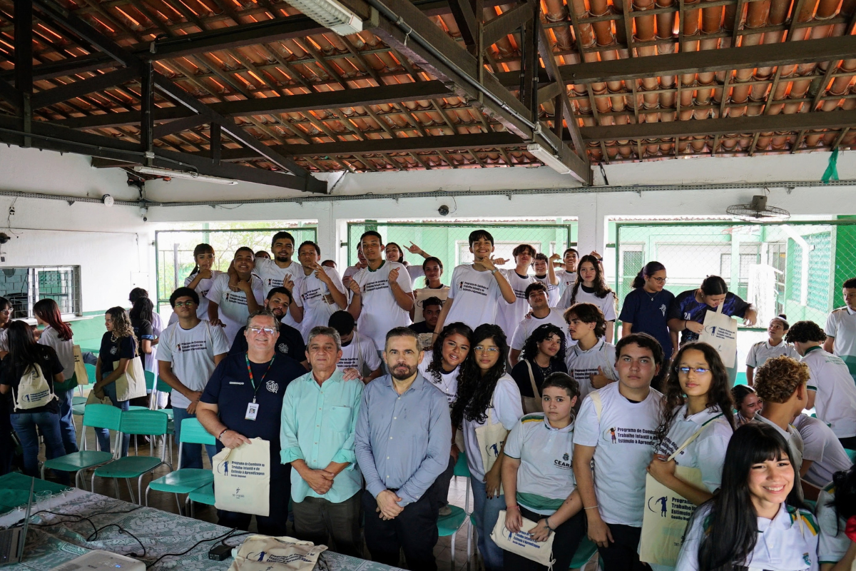 Fotografia de um grupo de jovens e adultos reunidos em um espaço coberto, com várias pessoas usando camisetas brancas e outras vestindo roupas escuras. Na parte frontal, três homens adultos aparecem lado a lado, acompanhados de outros participantes. Ao fundo, há cadeiras, janelas e uma quadra esportiva, enquanto parte do grupo segura bolsas de tecido.