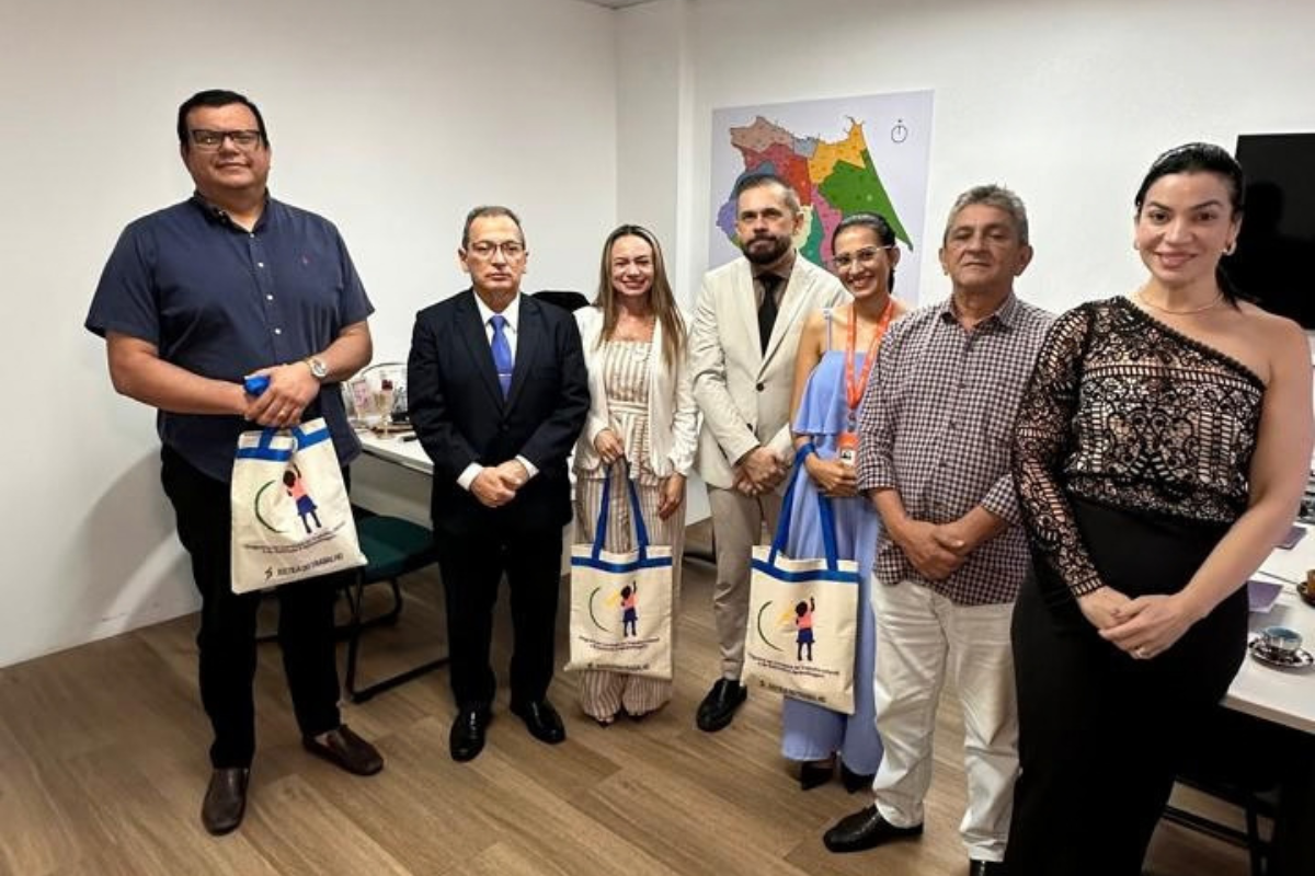Fotografia de sete pessoas, entre homens e mulheres, posando lado a lado em uma sala de escritório. Algumas vestem roupas sociais e outras, trajes casuais. Três delas seguram sacolas de tecido com ilustrações. Ao fundo, há mesas, cadeiras, computadores e um mapa colorido na parede.